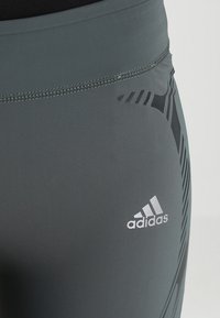 Szare legginsy sportowe o gładkiej fakturze, z czarnym wzorem graficznym po boku i białym logo Adidas na dolnej części nogawki.