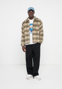 Plaid, beige and green wool jacket with a zipper, worn over a graphic white t-shirt. Paired with black cargo pants and light sneakers.