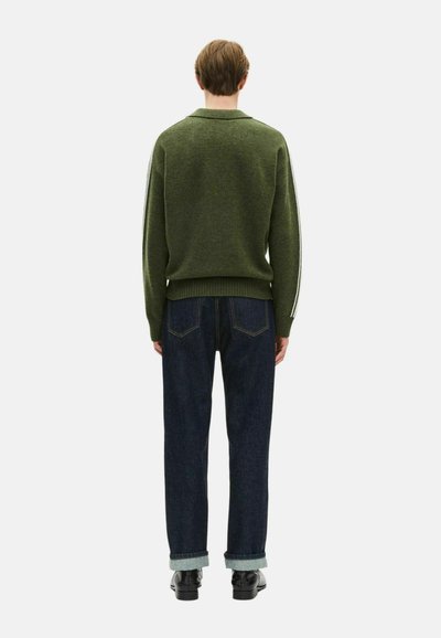 Una persona se encuentra de espaldas, vistiendo un suéter verde olivo con rayas blancas en las mangas, jeans oscuros arremangados y zapatos negros, sobre un fondo blanco.