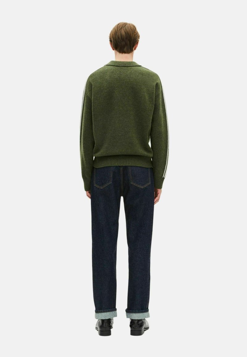 Une personne se tient de dos, portant un pull vert olive avec des rayures blanches sur les manches, un jean noir retroussé et des chaussures noires, sur un fond blanc.