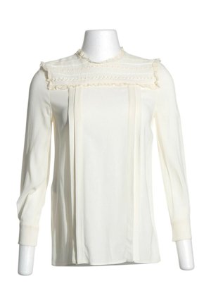 Blouse crème à manches longues avec col haut, bordure à volants et broderie horizontale décorative sur la poitrine.