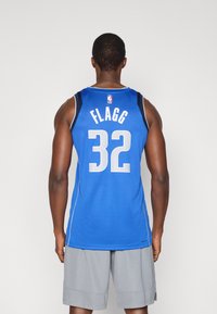 Μπλε φανέλα μπάσκετ με λευκές λεπτομέρειες, με το όνομα "FLAGG" και τον αριθμό "32" στην πλάτη. Φορεμένη με γκρι σορτς.