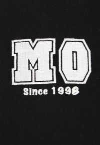 Fekete szövet, amelyen fehér hímzett "MO" betűk láthatók, alattuk "Since 1998" egyszerű, vastag betűtípussal. Puha tapintású, további minták nélkül.