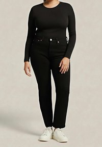 Femme portant un haut noir ajusté à manches longues, un jean noir taille haute et des baskets blanches, debout devant un fond uni.