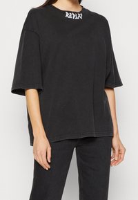 Camiseta negra oversize de algodón con cuello redondo, mangas cortas y logo de la marca en blanco contrastante en el cuello. Textura simple.