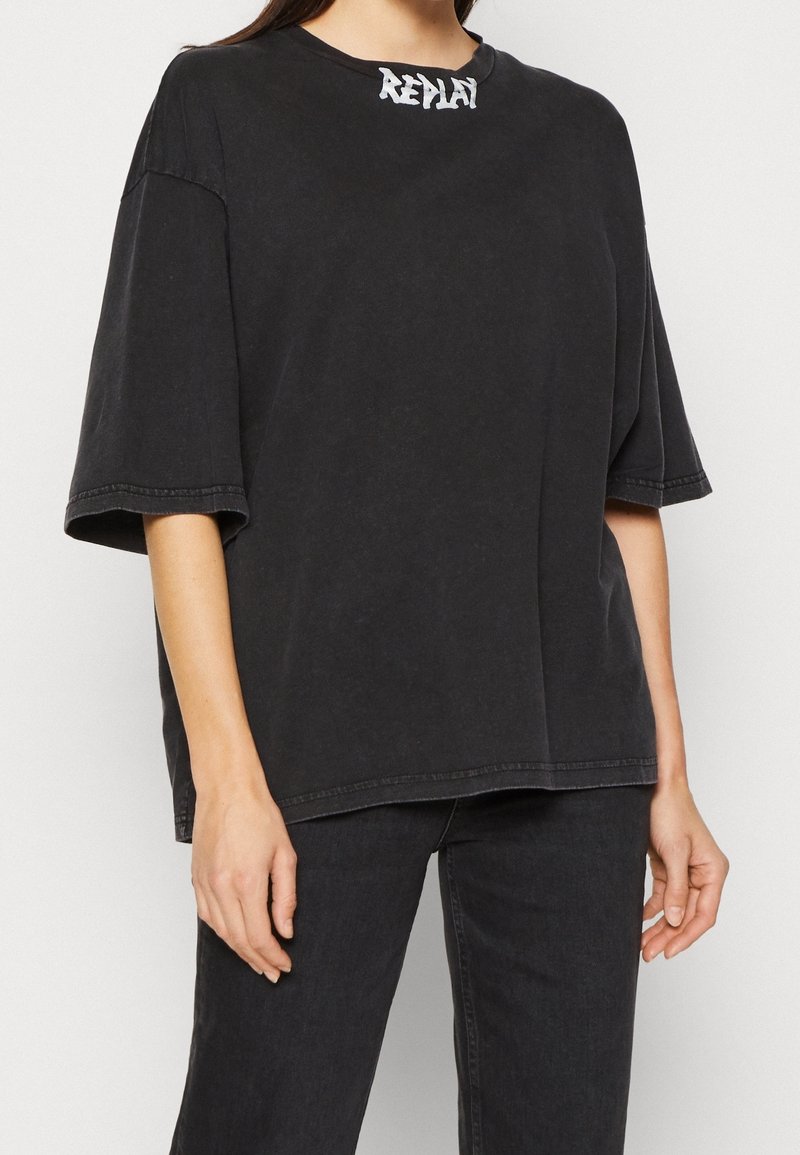 Camiseta negra oversize de algodón con cuello redondo, mangas cortas y logo de la marca en blanco contrastante en el cuello. Textura simple.
