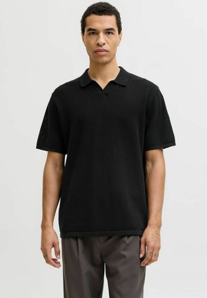 Mann trägt ein locker sitzendes, schwarzes, gestricktes Polohemd mit kurzen Ärmeln und graue Hosen, steht vor einem schlichten, hellen Hintergrund.