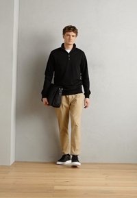 Suéter negro de manga larga con cuello con cremallera, chinos beige, zapatillas negras y un bolso bandolera negro, de pie contra una pared clara.
