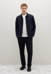 Camicia blu navy con bottoni sopra una t-shirt bianca, abbinata a pantaloni neri e sneaker nere. Modello in piedi con le mani in tasca. Sfondo semplice.