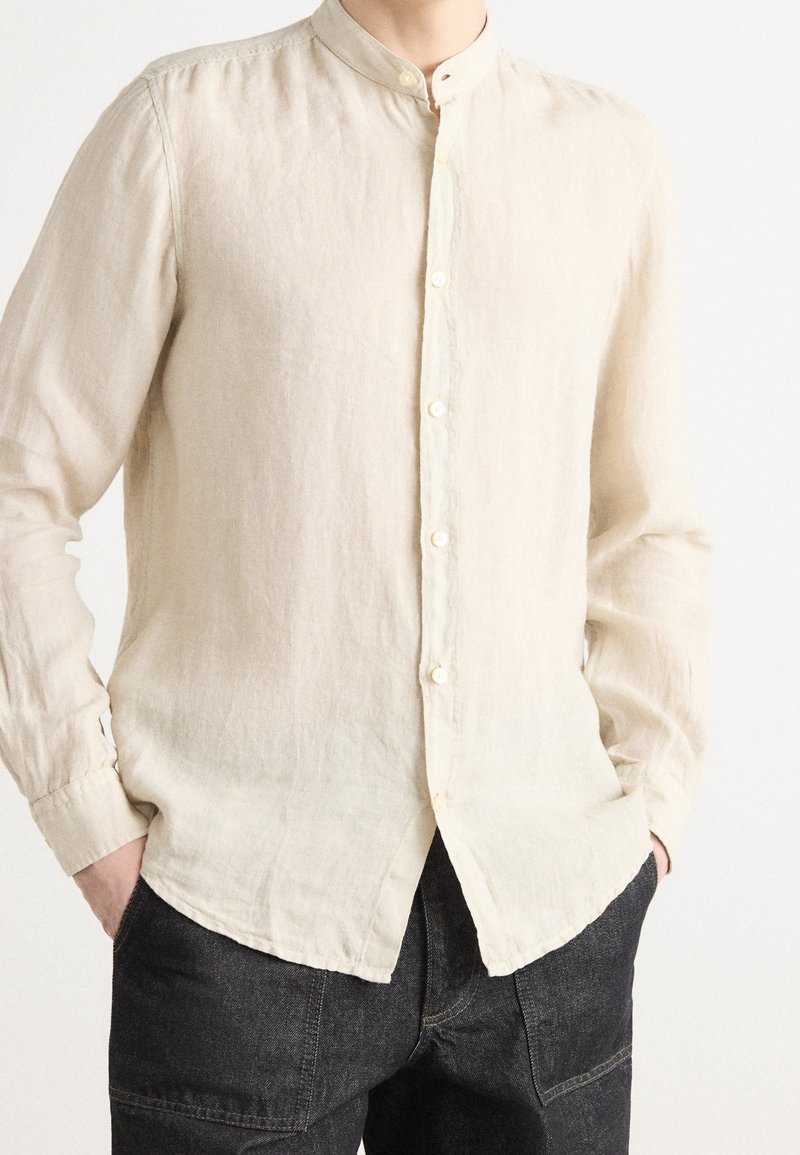 Homme portant une chemise crème à manches longues boutonnée avec col mandarin et pantalon foncé, mains dans les poches.