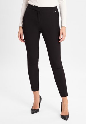 Pantalons noirs slim-fit en matériau extensible. Présentent un devant plat, des passants de ceinture et un petit accent logo. Stylés avec des talons aigus.