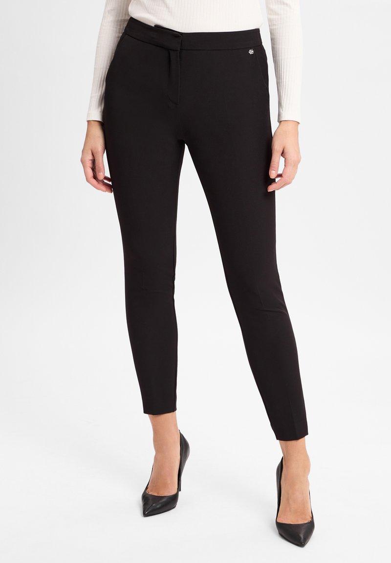 Pantaloni neri slim fit realizzati in materiale elasticizzato. Presentano una vita piatta, passanti per cintura e un piccolo logo decorativo. Abbinati con tacchi a punta.