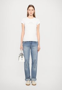 T-shirt bianco in cotone con logo, jeans in denim azzurro chiaro con dettagli decorati, borsa bianca e sneakers beige. Outfit casual.