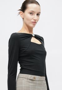 Νεαρή γυναίκα με μαλλιά χτενισμένα προς τα πίσω, που φοράει μαύρο μακρυμάνικο μπλουζάκι με κόπιτσα από στρας και γκρι καρό παντελόνι με ζώνη.