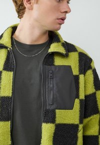 Fleece jas met een zwart en limegroen ruitpatroon, voorzien van een ritszak op de borst, gedragen over een donkergroen shirt.