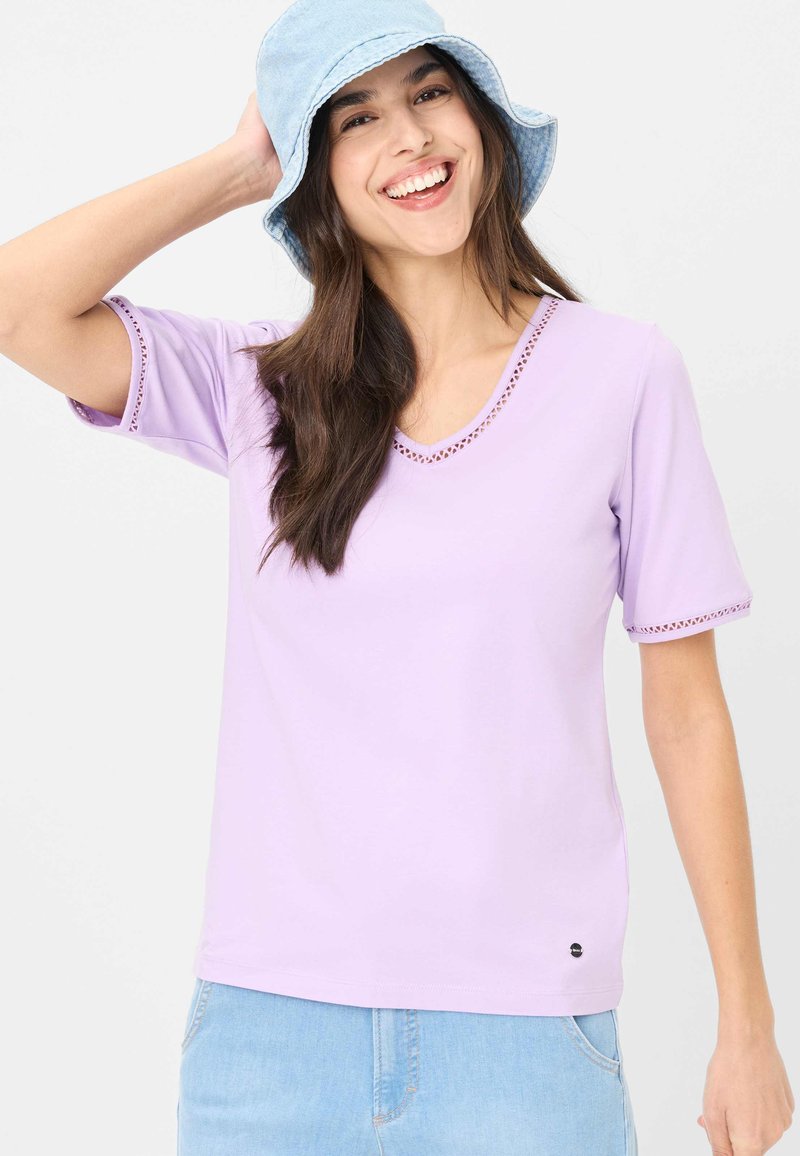 Lyse lilla kortærmet T-shirt med V-hals og blonder trim på ærmer og halsudskæring, lavet af et glat stof. Parret med en denim bucket hat.