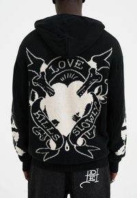 Hoodie en tricot noir avec un grand motif en forme de cœur blanc et le texte "L'AMOUR TUE SLOWLY." Comprend des détails complexes sur les manches.