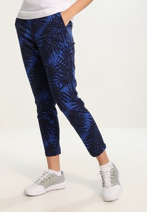 Osoba ubrana w niebiesko-czarne, cienkie spodnie w liście, nosząca białe i szare sneakersy, stojąca z jedną ręką w kieszeni.