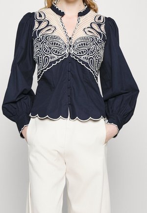 Blouse marine avec détails en dentelle blanche et bords festonnés, manches longues bouffantes, devant à boutons, associée à un pantalon blanc taille haute.
