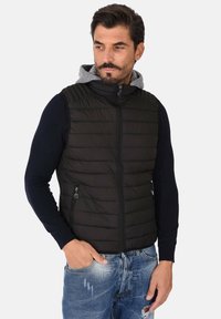 Gilet nero senza maniche con design trapuntato, zip davanti e due tasche laterali, indossato sopra una camicia a maniche lunghe blu navy. Jeans in denim casual.