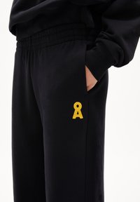 Schwarze Sweatpants mit einem elastischen Bund, seitlichen Taschen und einem kleinen gelben gestickten Logo am linken Oberschenkel. Glatte Stofftextur.