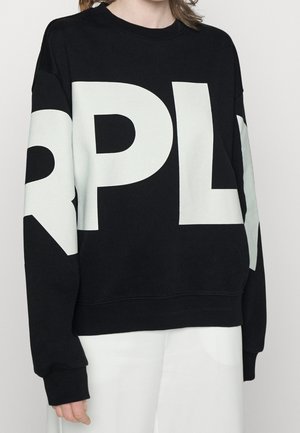 Person iført en sort sweatshirt med store hvide bogstaver "RPL" over front og ærmer, parret med hvide bukser.