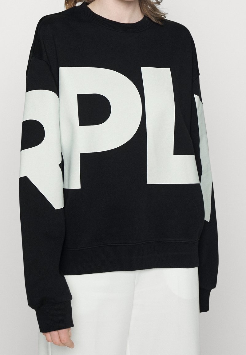 Personne portant un sweat-shirt noir avec de grandes lettres blanches "RPL" sur le devant et les manches, associé à un pantalon blanc.
