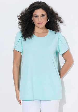 Kvinde med krøllet mørkt hår iført sølv øreringe i ring og en lyseblå kortærmet top, står foran en hvid baggrund.