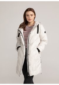 Witte gewatteerde lange jas met een capuchon, voorzien van diagonale stiksels, zwarte accenten en zijzakken, gelaagd over een gestreept shirt.