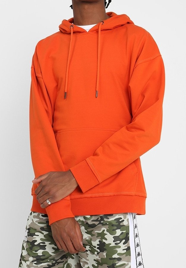 Personne portant un sweat à capuche orange vif et un short camouflage vert avec des rayures blanches sur les côtés, debout les bras croisés.