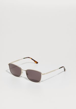Ochelari de soare cu ramă din metal de culoare aurie și lentile închise. Brațele subțiri au un model de țestoasă și un accent verde lângă balama.