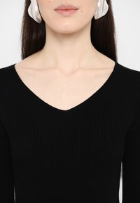 Musta ribbed-neulospaita syvällä V-pääntiellä, joka korostaa vartalon muotoa ja on miellyttävän sileä. Hopeiset näyttävät korvakorut täydentävät asun saraa.