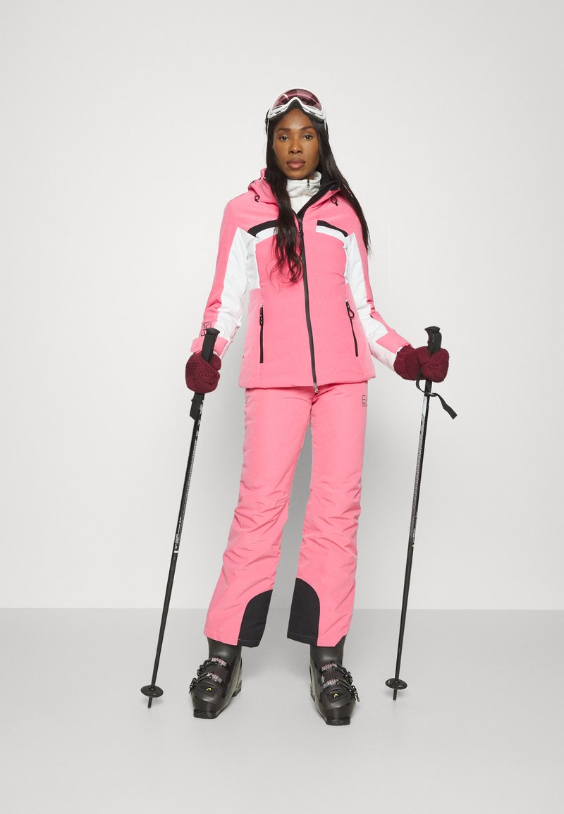 Roze en witte skijack met ritszakken en zwarte accenten, gecombineerd met bijpassende roze skibroek. Draagt skischoenen en houdt stokken vast.