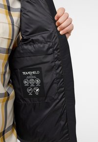 Schwarze, isolierte Jackeninnenseite mit einem Etikett, das die Texashield-Marke zeigt, sowie Symbole für winddicht und wasserabweisend und Details zum natürlichen Daunenmaterial.