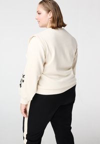 Lichtbeige sweatshirt met geplooide mouwen en zwarte bloemrijke borduursels op de linker mouw, gecombineerd met zwarte broek met een zijstreep.