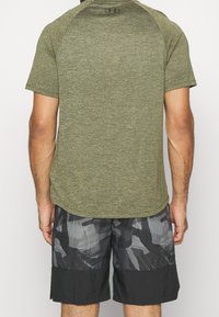 Olivově zelené sportovní tričko s krátkým rukávem a zakřiveným lemem; kombinováno s šortkami v černobílém maskáčovém vzoru, které mají elastický pas.