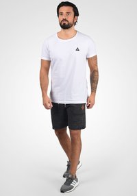 Camiseta blanca de algodón con un pequeño logotipo triangular negro; combinada con pantalones cortos gris oscuro con un patrón texturizado y un acento en forma de diamante marrón.