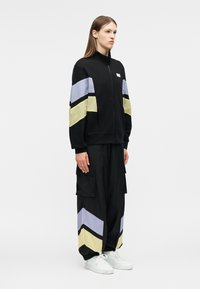 Femme debout portant un survêtement noir avec des rayures en chevron jaune et violet clair sur les manches et le pantalon, associée à des baskets blanches.