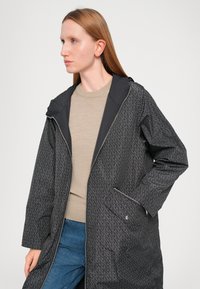 Lange zwarte jas met geometrisch patroon, voorzien van een capuchon, een ritssluiting aan de voorkant en twee zijzakken. Draagt over een beige top en denim.