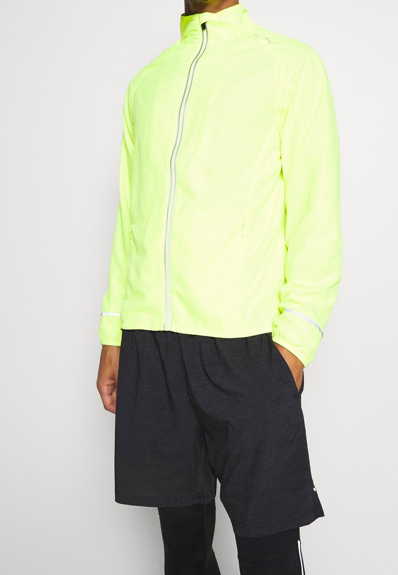 Homme portant une veste zippée jaune néon avec des détails réfléchissants, un short noir et des leggings noirs sur un fond uni.