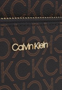 Černá texturovaná peněženka s opakujícím se hnědým vzorem loga, s výrazným zlatým emblémem "Calvin Klein". Součástí je zlatý zip.