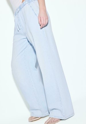 Personne portant un pantalon large bleu clair avec une ceinture élastique et des chaussures blanches en mesh pointues, debout sur fond blanc.