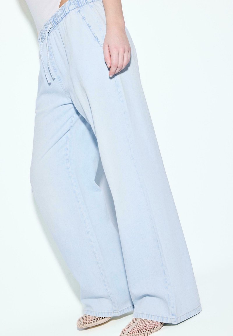 Personne portant un pantalon large bleu clair avec une ceinture élastique et des chaussures blanches en mesh pointues, debout sur fond blanc.