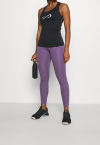 Débardeur de sport noir avec logo blanc ; leggings ajustés violets ; chaussures de sport noires ; tenant une bouteille d'eau noire. Texture lisse, design moderne.