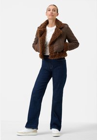 Brun shearling-krage läderjacka, vit texturerad tröja, mörkblå vida jeans och vita sneakers med beige accenter.