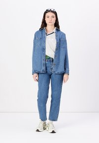 Giacca di jeans con due tasche sul petto, chiusura con bottoni; abbinata a una camicia grigia e jeans blu a vita alta. Sneakers di colore chiaro.