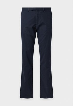 Un homme porte un pull torsadé couleur crème sur une chemise à carreaux, un pantalon bleu marine, des baskets vertes et une casquette sombre, sur un fond gris uni.
