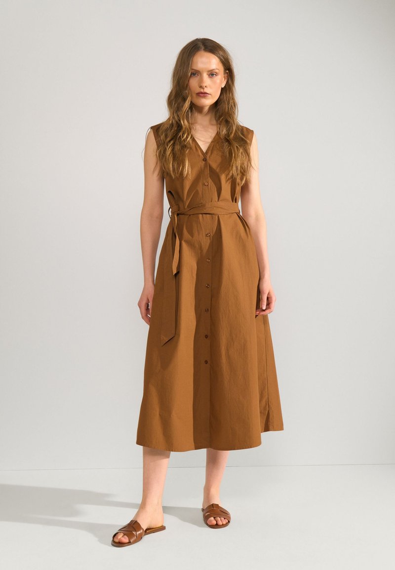 Femme aux longs cheveux portant une robe midi marron sans manches boutonnée avec une ceinture, debout droite avec des sandales plates marron.