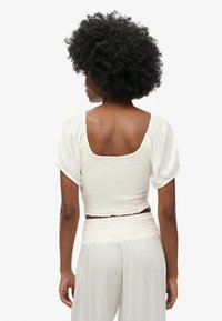 Top blanc court avec des manches bouffantes, présentant un design texturé et froncé. L'arrière a un large décolleté carré et une coupe décontractée.