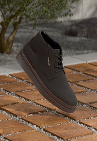 Dark brown GANT leather sneaker with laces, floating above wooden square tiles set in gravel outdoors.